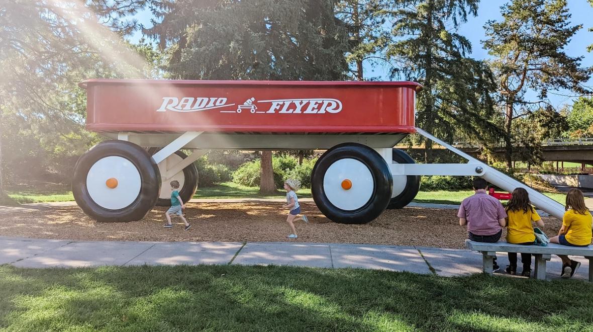 Young kids run in front of a giant red Radio Flyer wagon sculpture in Spokane's Riverfront Park. Kids can slide down the handle.