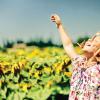 A young girl standing in a sunflower field with her arms raised above her head and her face looking up at the sun with her eyes closed and smiling