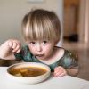 little boy sitting at a table eating a bowl of soup