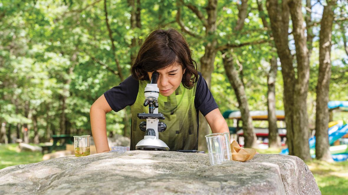 boy bent over a microscope resting on a large rock outside