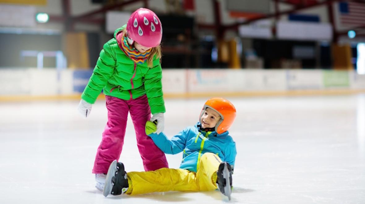 Two children ice skating indoors, one sitting on the ice smiling while the other helps them up