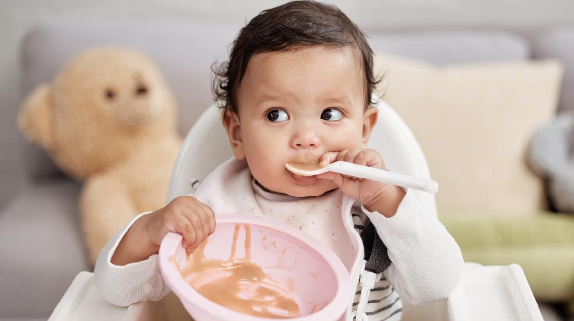 Baby in a highchair holding an empty bowl and spoon as a way to wean baby to solid foods