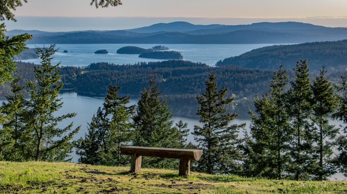 Empty wooden bench with panoramic view of many islands in Puget Sound, sunset in distance, slight haze to sky, from Turtleback Mountain on Orcas Island