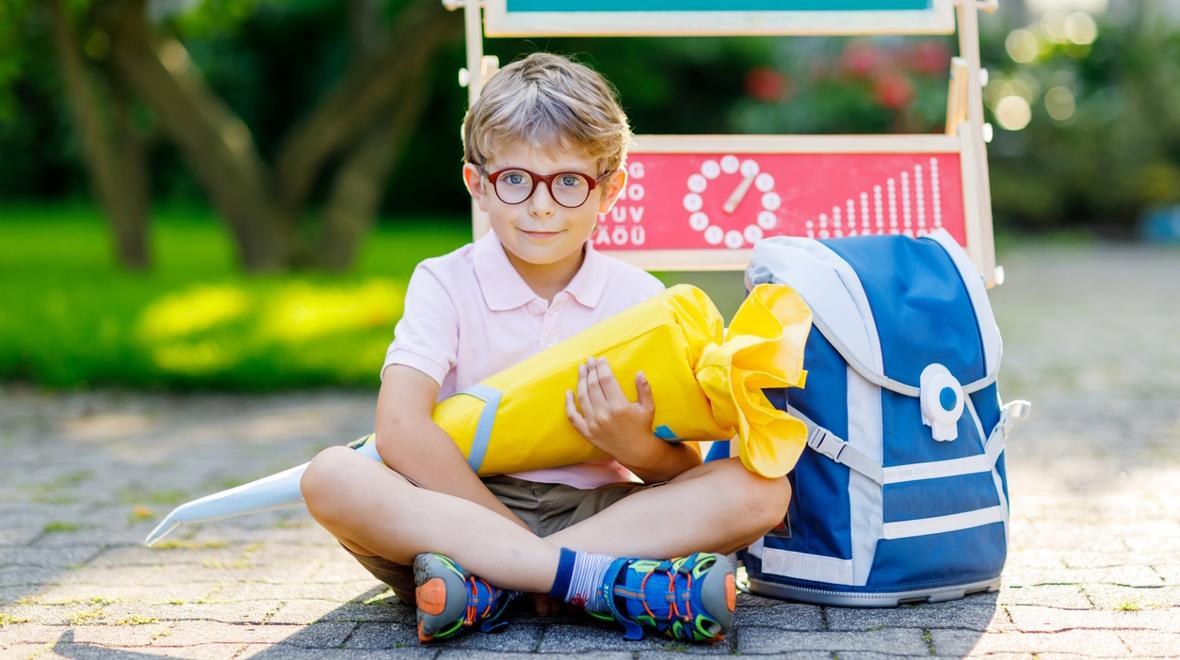 boy sitting on the sidewalk with a blue backpack holding a yellow back-to-school Schultuete 