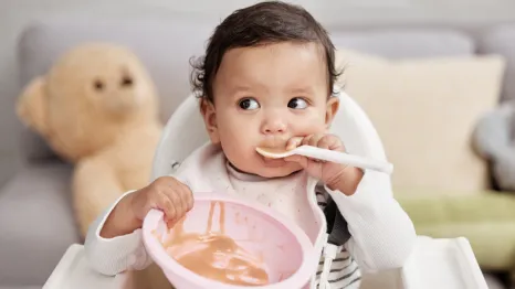 Baby in a highchair holding an empty bowl and spoon as a signal for baby led weaning