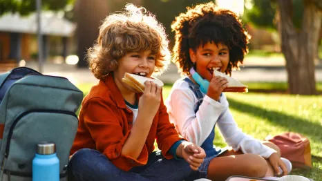 two kids sitting in the grass eating a healthy lunch at summer camp
