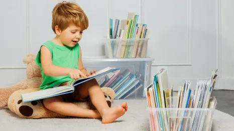 Young boy sitting in a play room with a stuffed bear with plastic containers of books