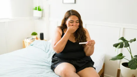 a surprised woman sitting on a bed looking at a pregnancy test 