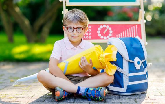 boy sitting on the sidewalk with a blue backpack holding a yellow back-to-school Schultuete 