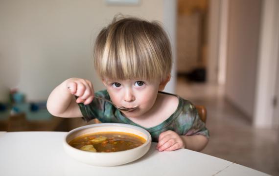 little boy sitting at a table eating a bowl of soup