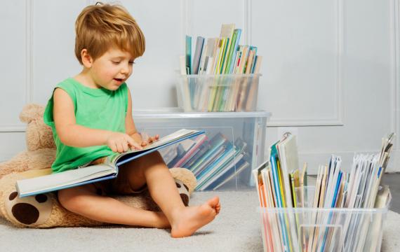 Young boy sitting in a play room with a stuffed bear with plastic containers of books