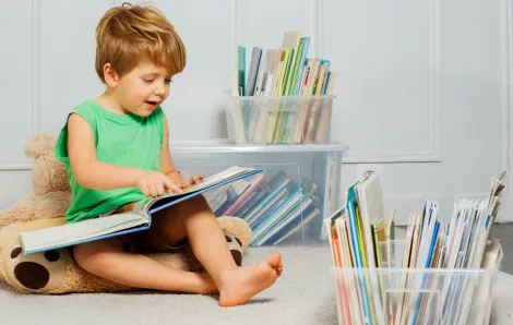 Young boy sitting in a play room with a stuffed bear with plastic containers of books