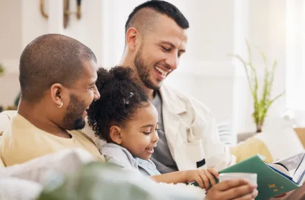 Two dads sitting on the couch with their daughter reading a book with LGBTQ characters