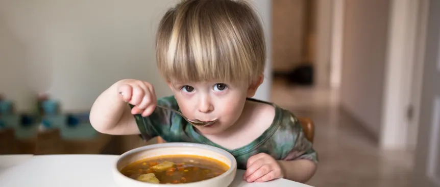 little boy sitting at a table eating a bowl of soup