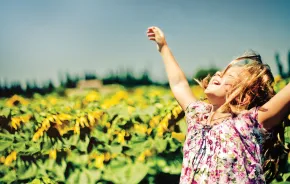 A young girl standing in a sunflower field with her arms raised above her head and her face looking up at the sun with her eyes closed and smiling