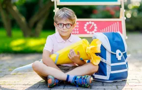 boy sitting on the sidewalk with a blue backpack holding a yellow back-to-school Schultuete 