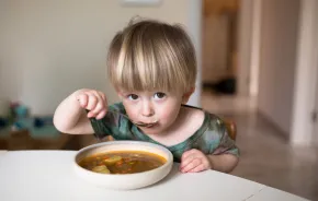 little boy sitting at a table eating a bowl of soup