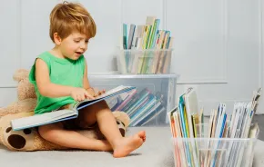 Young boy sitting in a play room with a stuffed bear with plastic containers of books